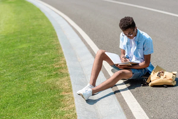 Blick aus der Vogelperspektive auf einen afrikanisch-amerikanischen Studenten, der auf einem Skateboard sitzt und im Park ein Buch liest — Stockfoto