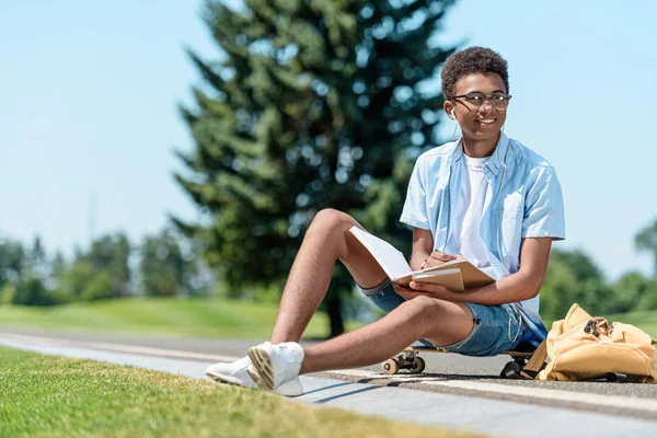 Sonriente afroamericano adolescente sentado en monopatín y lectura libro en parque - foto de stock