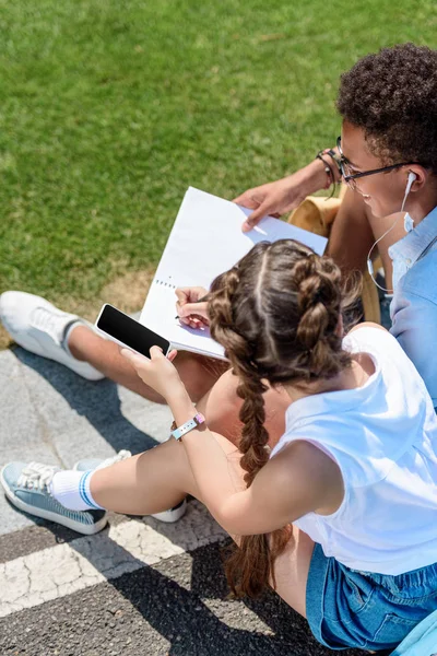 Vista de ángulo alto de adolescente multiétnico y niña usando teléfono inteligente y estudiando juntos en el parque - foto de stock