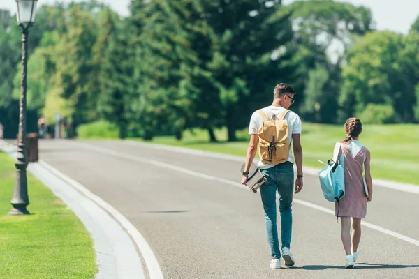 Back view of teenage boy and girl with backpacks and skateboard walking together in park — Stock Photo