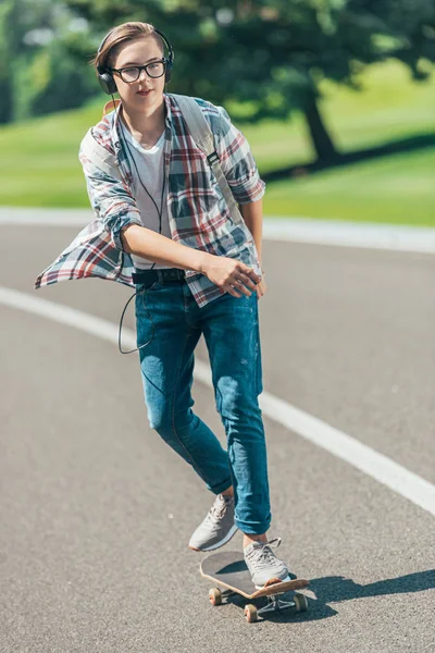 Студентка-підліток в навушниках їде скейтборд і дивиться в парк — стокове фото