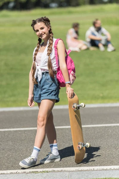 Красива дівчина-підліток з рюкзаком, що стоїть зі скейтбордом і посміхається на камеру в парку — стокове фото