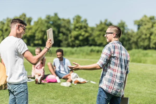 Happy teenage boys smiling each other and holding book while friends studying behind in park — Stock Photo