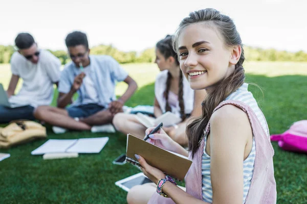 Happy teenage girl taking notes and smiling at camera while studying with friends in park — Stock Photo