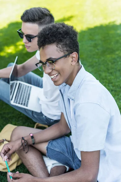 Blick aus der Vogelperspektive auf multiethnische Teenager, die beim gemeinsamen Lernen im Gras lächeln und Laptop benutzen — Stockfoto