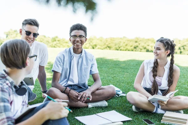 Estudiantes adolescentes multiétnicos felices sentados en la hierba y estudiando juntos en el parque - foto de stock