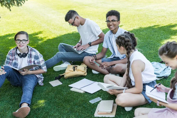 Vista ad alto angolo di sorridenti adolescenti multietnici seduti sull'erba e che studiano nel parco — Foto stock