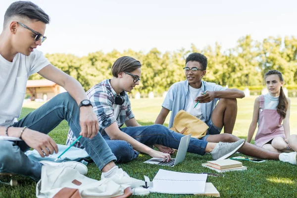 Adolescentes multiétnicos felices sentados y hablando mientras estudian juntos en el parque - foto de stock