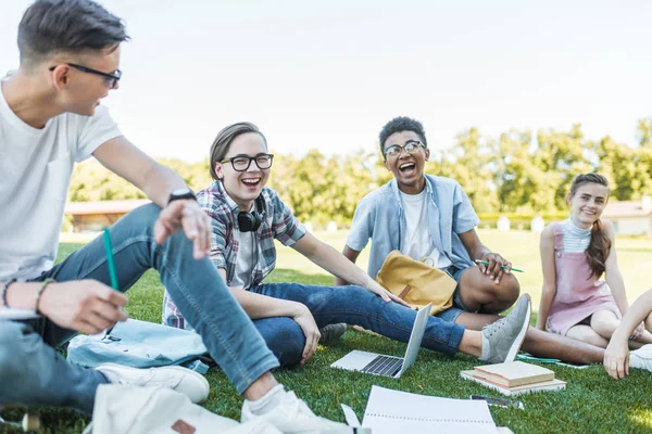 Adolescentes multiétnicos felices sentados y riendo mientras estudian juntos en el parque - foto de stock