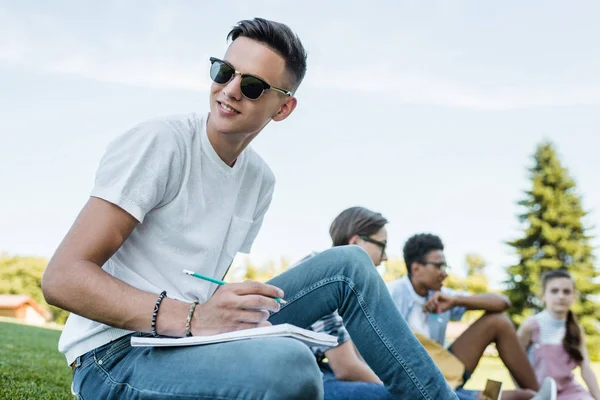 Sorridente adolescente con gli occhiali da sole prendere appunti e distogliere lo sguardo mentre studia con gli amici nel parco — Foto stock