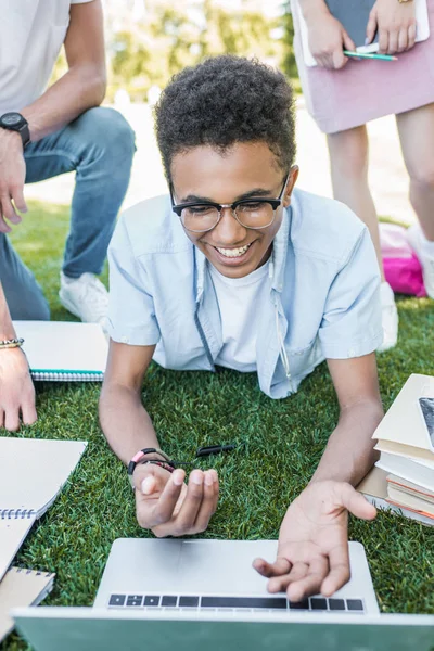 Niño afroamericano sonriente usando portátil mientras estudia con amigos en el parque - foto de stock