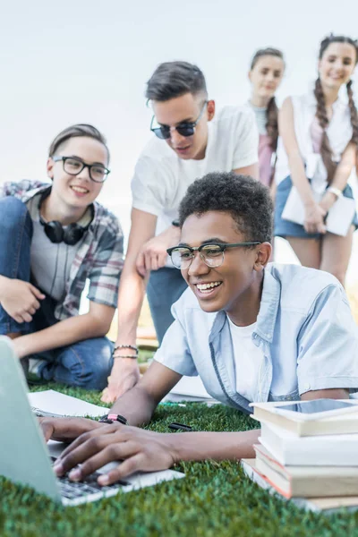 Amigos adolescentes multiétnicos felices usando el ordenador portátil y estudiando en el parque - foto de stock