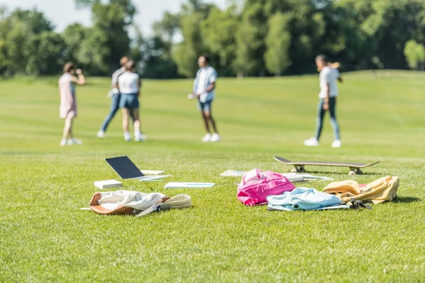 Computer portatile, libri, zaini e skateboard su erba e adolescenti che giocano dietro nel parco — Foto stock