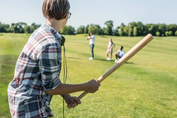 Vista lateral del adolescente sosteniendo bate de béisbol y jugando con amigos en el parque - foto de stock
