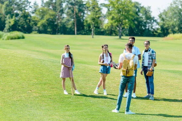 Fröhliche multiethnische Teenager-Klassenkameraden mit Rucksäcken spielen Baseball im Park — Stockfoto