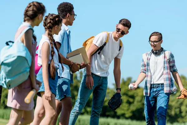 Smiling multiethnic friends with books and backpacks looking at classmates with baseball ball and gloves in park — Stock Photo