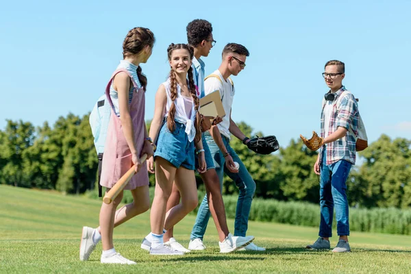 Amigos adolescentes multiétnicos felices con libros, mochilas y guantes de béisbol caminando juntos en el parque - foto de stock