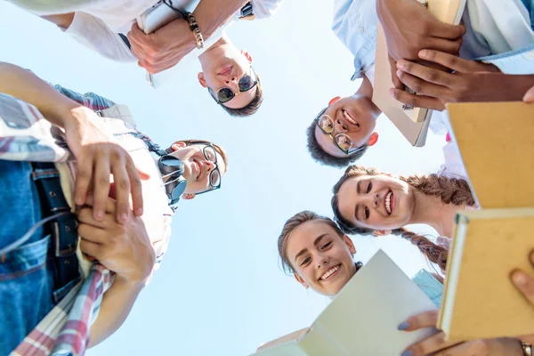 Visão inferior de adolescentes felizes amigos multiétnicos segurando livros e de pé juntos contra o céu azul — Fotografia de Stock