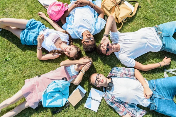 Вид зверху багатоетнічних однокласників-підлітків з книгами та рюкзаками, що лежать разом на зеленому лузі — стокове фото