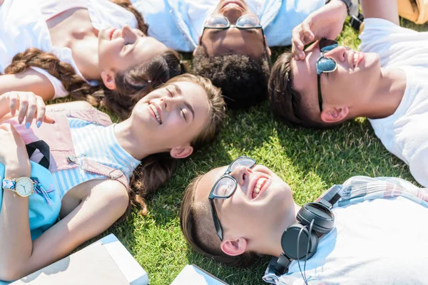 Високий кут зору щасливих багатоетнічних друзів-підлітків, що лежать разом на зеленому лузі — стокове фото