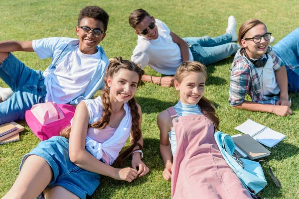 Glückliche Teenager-Freunde mit Büchern und Rucksäcken, die auf der Wiese sitzen und in die Kamera lächeln — Stockfoto