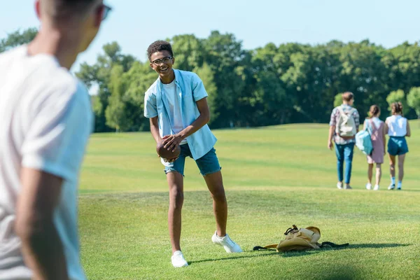 Enfoque selectivo de amigos adolescentes multiétnicos jugando con pelota de rugby en el parque - foto de stock