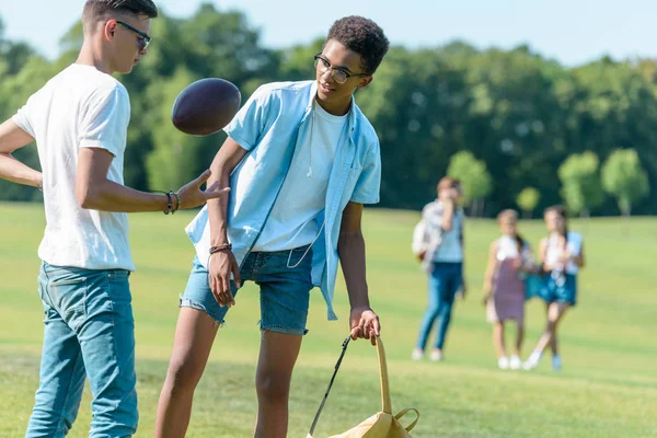 Adolescentes amigos multiétnicos jugando con pelota de rugby mientras sus compañeros de clase caminan detrás en el parque - foto de stock