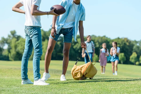 Recortado tiro de multiétnicos chicos jugando con pelota de rugby mientras compañeros de clase caminando detrás en parque - foto de stock