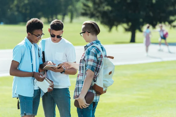 Adolescentes multiétnicos con libros y mochilas de pie y hablando juntos en el parque - foto de stock