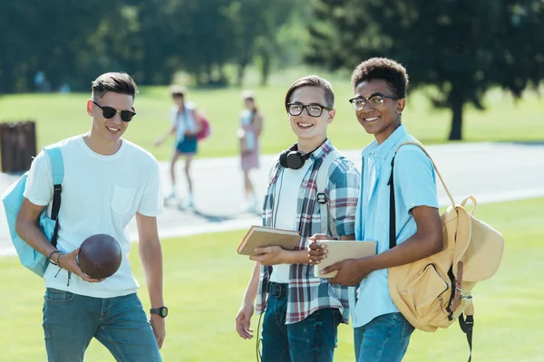 Adolescentes multiétnicos con libros y mochilas sonriendo a la cámara en el parque - foto de stock