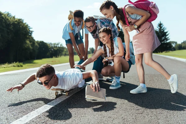 Happy teenage classmates looking at smiling boy lying on skateboard in park — Stock Photo