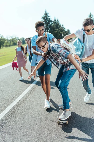 Happy multiethnic teenage classmates having fun with skateboard and walking together in park — Stock Photo