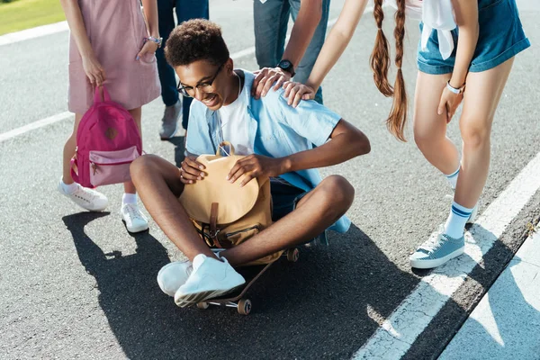 Ritagliato colpo di adolescenti in piedi vicino sorridente ragazzo africano americano tenendo lo zaino e seduto su skateboard — Foto stock