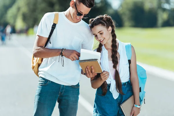 Happy teenagers with backpacks holding books while standing and smiling together in park — Stock Photo