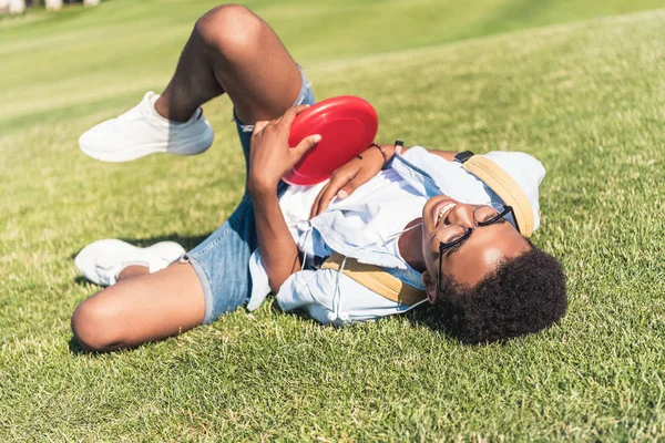 Feliz adolescente afro-americano segurando disco deitado e deitado na grama no parque — Fotografia de Stock