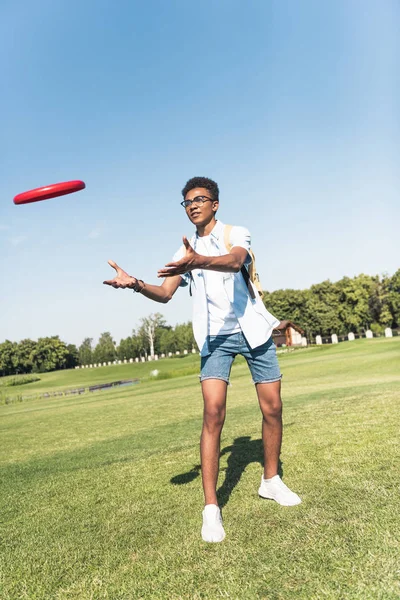 Visão de comprimento total do adolescente afro-americano com mochila brincando com disco voador no parque — Fotografia de Stock