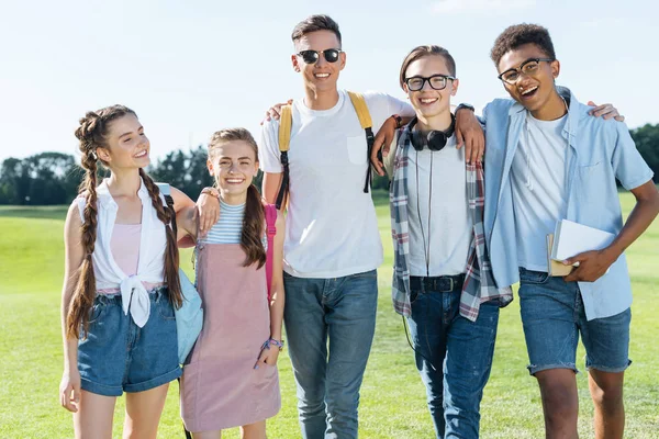 Feliz multiétnico adolescentes amigos de pie juntos y sonriendo a la cámara en el parque - foto de stock