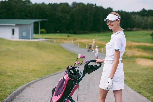 Selektiver Fokus einer lächelnden Golfspielerin in Mütze und weißem Polo mit Golfausrüstung auf dem Golfplatz — Stockfoto