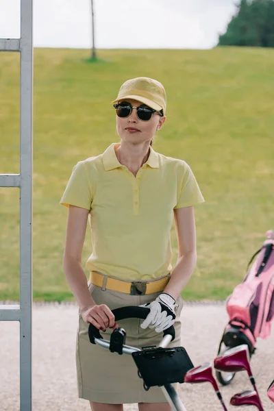 Портрет жіночого гравця в шапці і сонцезахисних окулярах, що стоять біля обладнання для гольфу на полі для гольфу — стокове фото