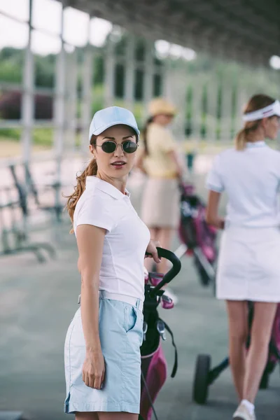 Selektiver Fokus der Frau mit Sonnenbrille und Golfausrüstung, die auf dem Golfplatz in die Kamera schaut — Stockfoto