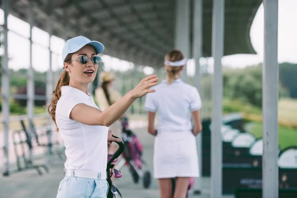 Foco seletivo da mulher em óculos de sol com equipamento de golfe apontando para longe no campo de golfe — Fotografia de Stock