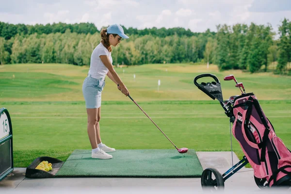 Vista lateral da mulher no pólo e boné jogando golfe no campo de golfe — Fotografia de Stock