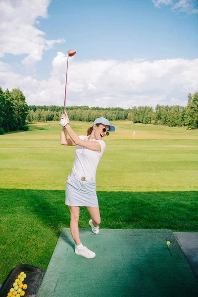 Вид збоку жінки в поло, сонцезахисні окуляри і кепка, граючи в гольф на полі для гольфу — стокове фото