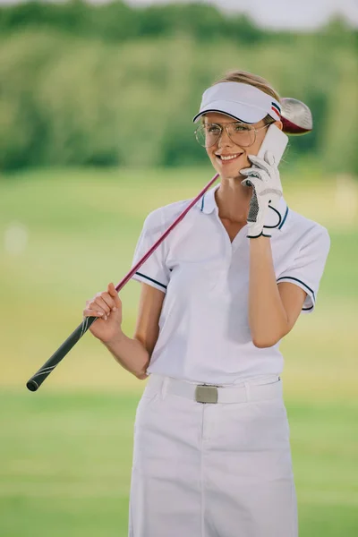 Portrait of smiling female golf player in polo and cap with golf club in hand talking on smartphone at golf course — Stock Photo