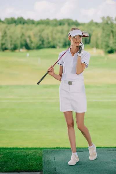 Гольфистка в поло и кепке с клюшкой для гольфа в руке разговаривает по смартфону на поле для гольфа — стоковое фото