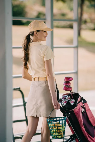 Side view of smiling woman in polo and cap carrying golf equipment at golf course — Stock Photo
