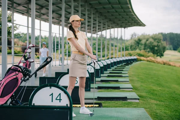 Pensive female golfer in polo and cap with golf club looking away at golf course — Stock Photo