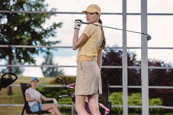 Enfoque selectivo de jugador de golf femenino con club de golf y amigo descansando detrás en el campo de golf - foto de stock