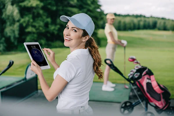 Selective focus of smiling woman with tablet and friend playing golf behind at golf course — Stock Photo