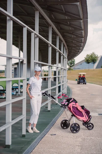 Smiling female golf player in while polo and cap with golf equipment at golf course — Stock Photo